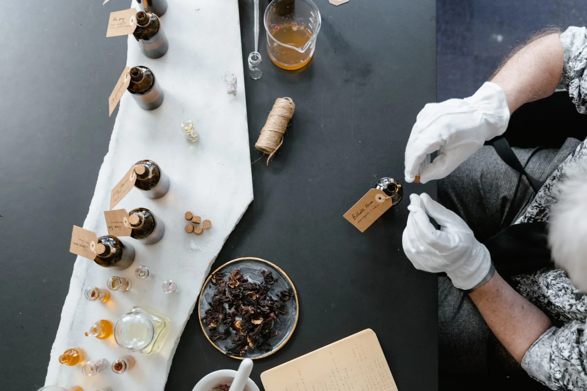 Overhead view of the bench: gloved hands, small bottles and a dish of dried petals