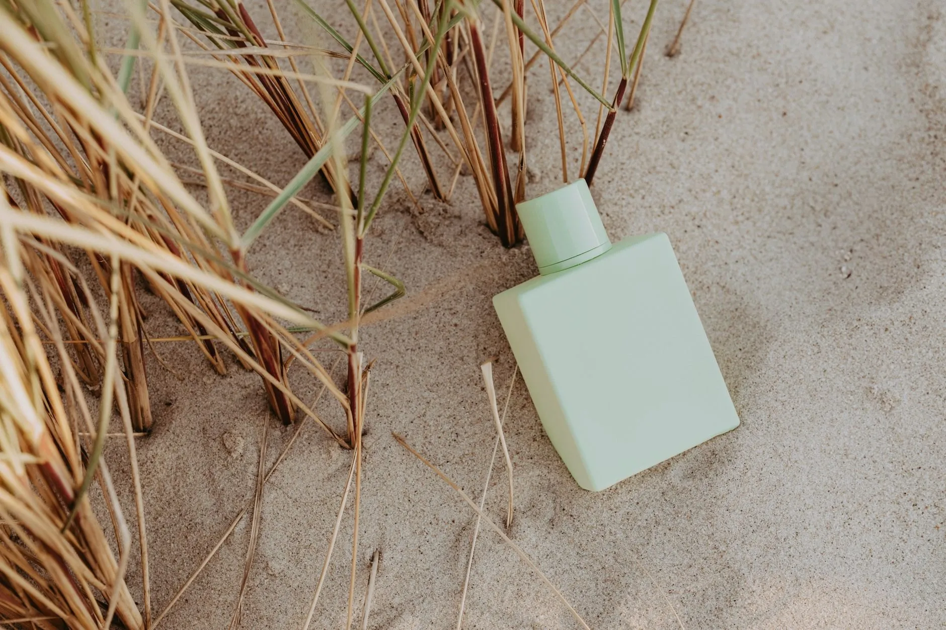 A sage-green Orrishaw bottle resting in pale sand among dry dune grass