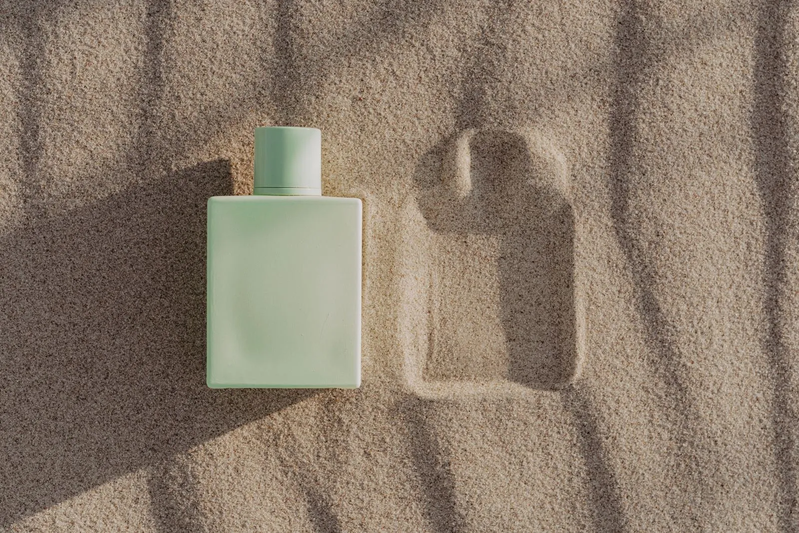 A sage-green Orrishaw bottle lying in sand beside the imprint it left